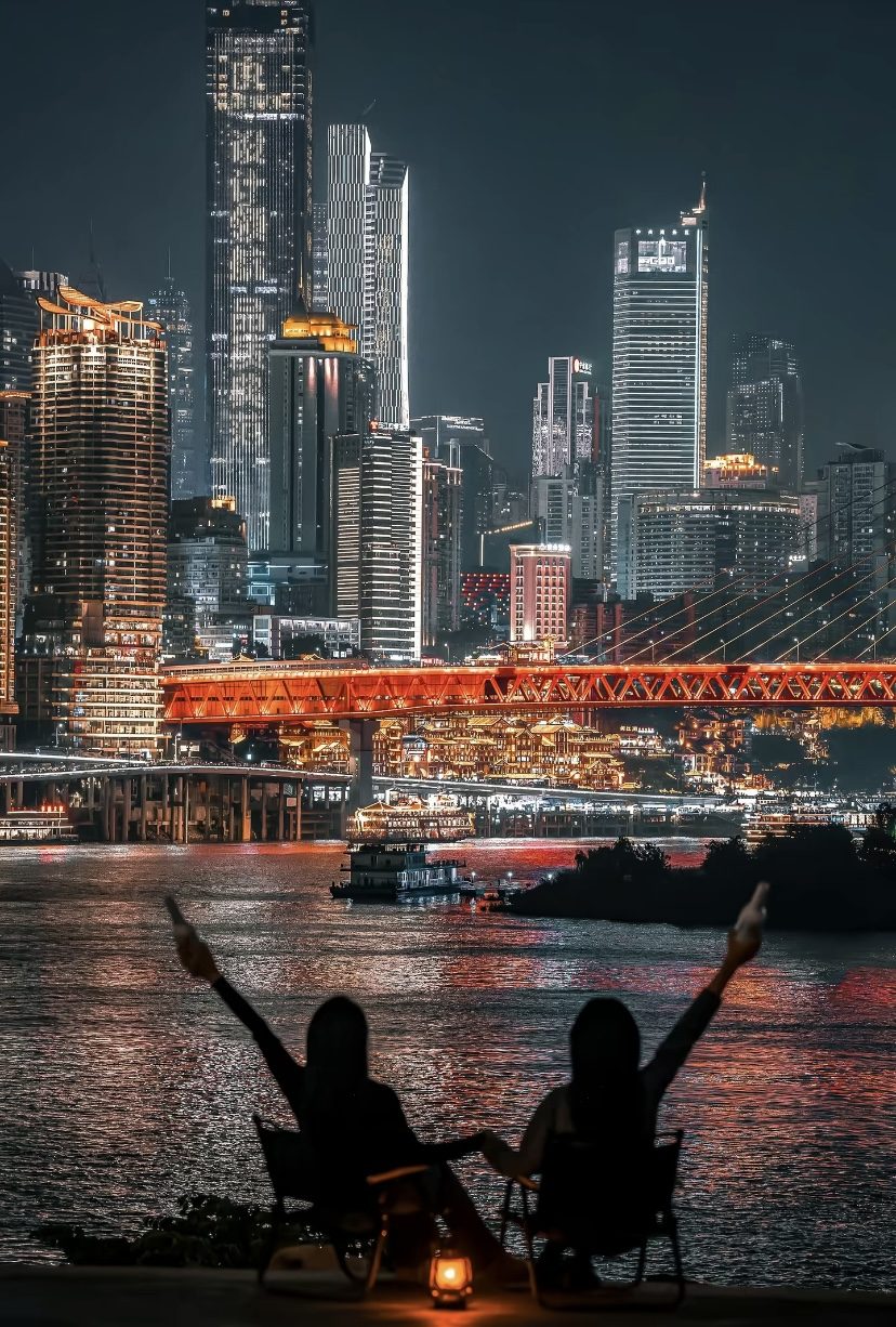 Two travelers enjoying the illuminated Chongqing skyline from a riverside viewpoint at night