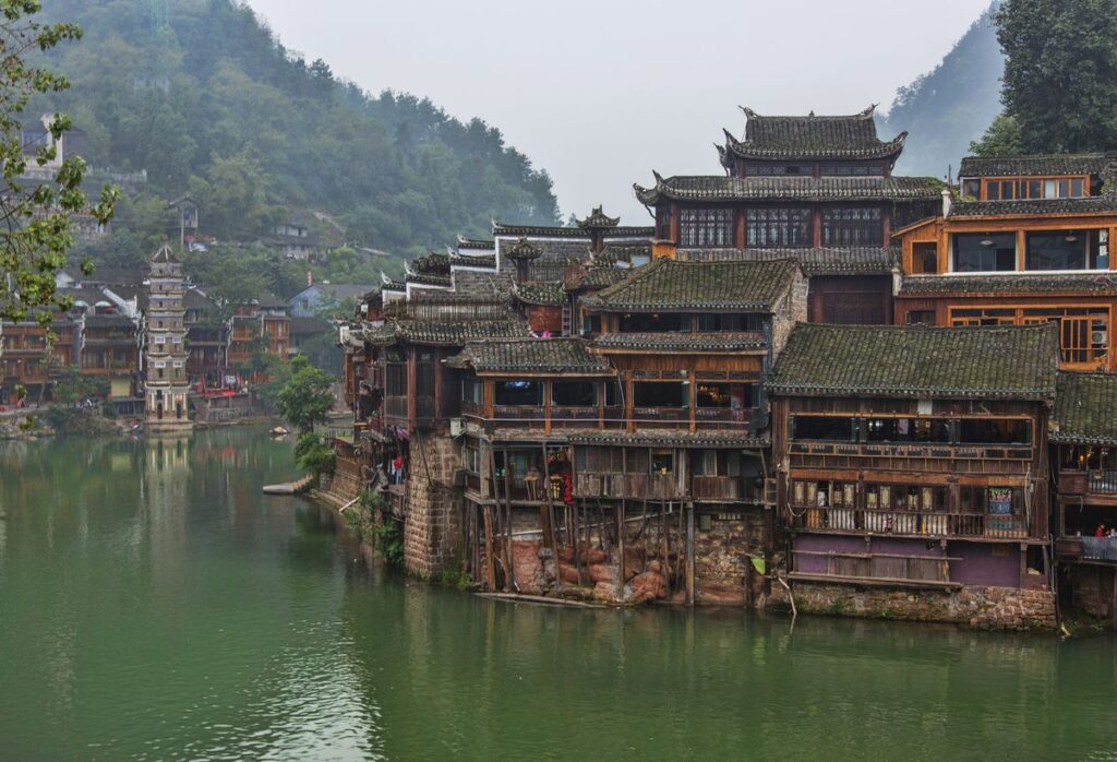 Wooden stilt houses along the green Tuojiang River in Fenghuang Ancient Town, with a pagoda in the distance