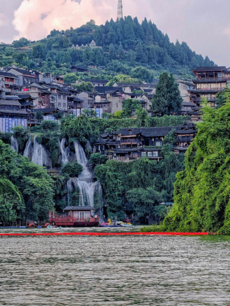 Furong Ancient Town and its waterfall seen from the Youshui River, with a sightseeing boat below