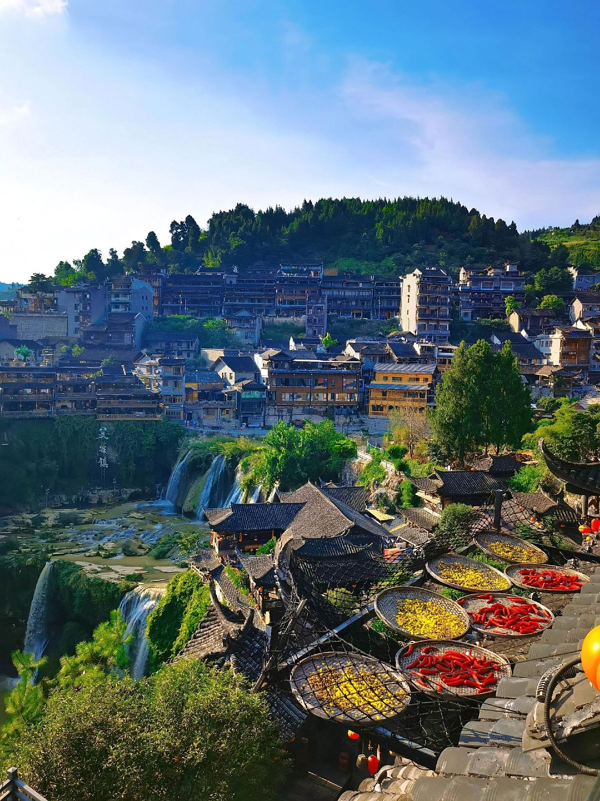 Furong Town's wooden houses on the cliff above the waterfall on a sunny day, with chillies drying on a rooftop