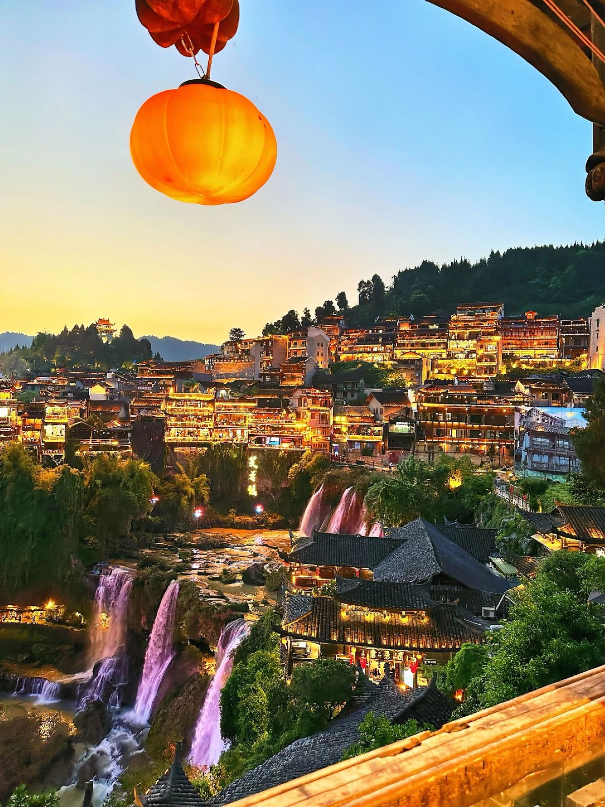 Furong Town at dusk, lit houses along the cliff and the waterfall below, seen from a balcony with a lantern