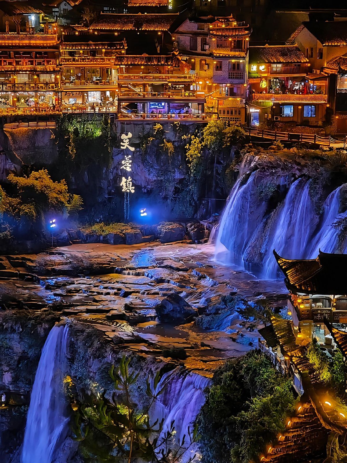 Furong Town waterfall lit at night below the illuminated houses and the Furong Town sign on the cliff