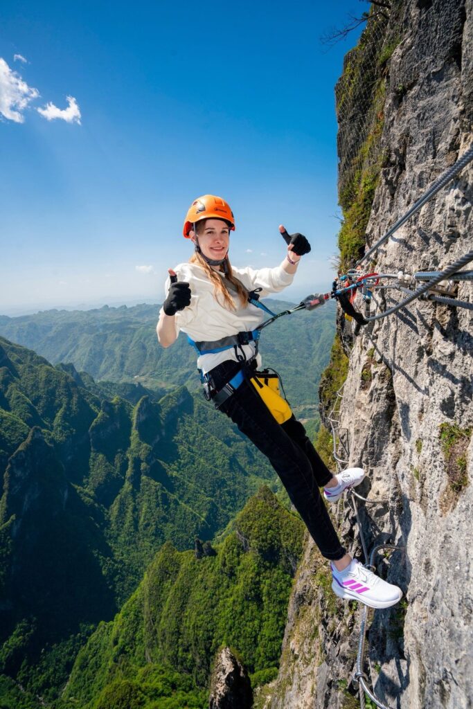 A climber in helmet and harness on the Qixing Mountain via ferrata, standing on steel rungs bolted to the cliff face above the valley