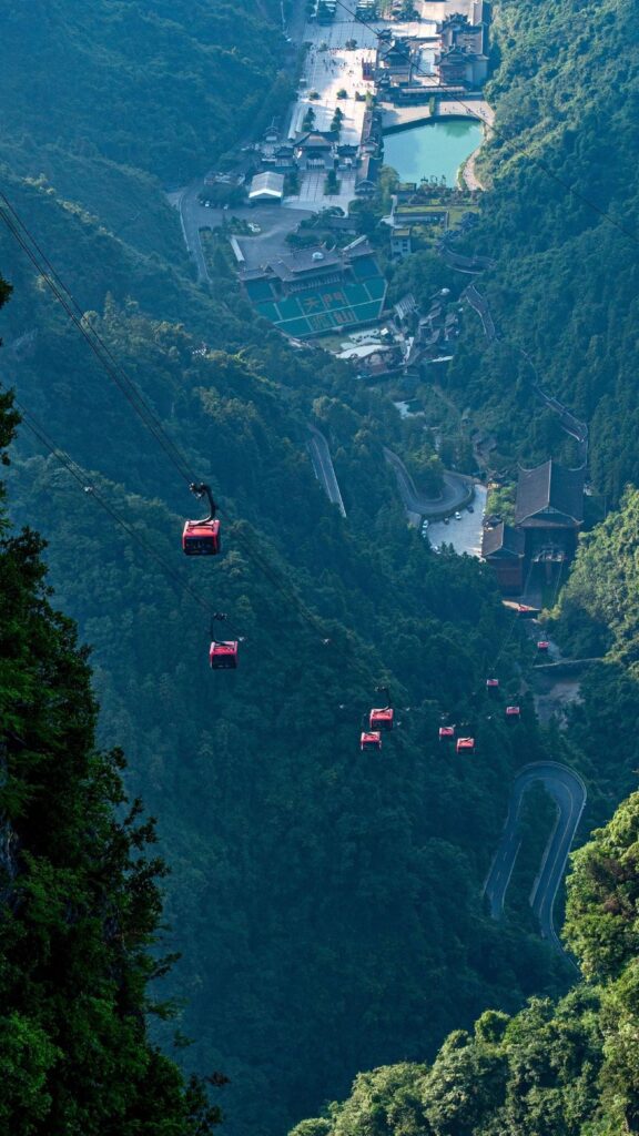 Red cabins of the Tianmen Cave express cable car crossing above the 99-bend road, with the theatre and lake at the mountain gate below