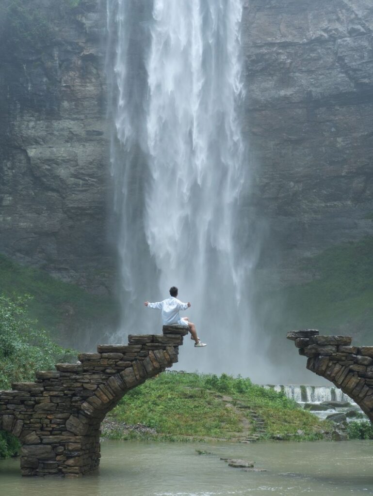 Liusha Waterfall pouring down a dark cliff in the Dehang Grand Canyon, Xiangxi, with a traveller sitting on an old broken stone arch bridge in front
