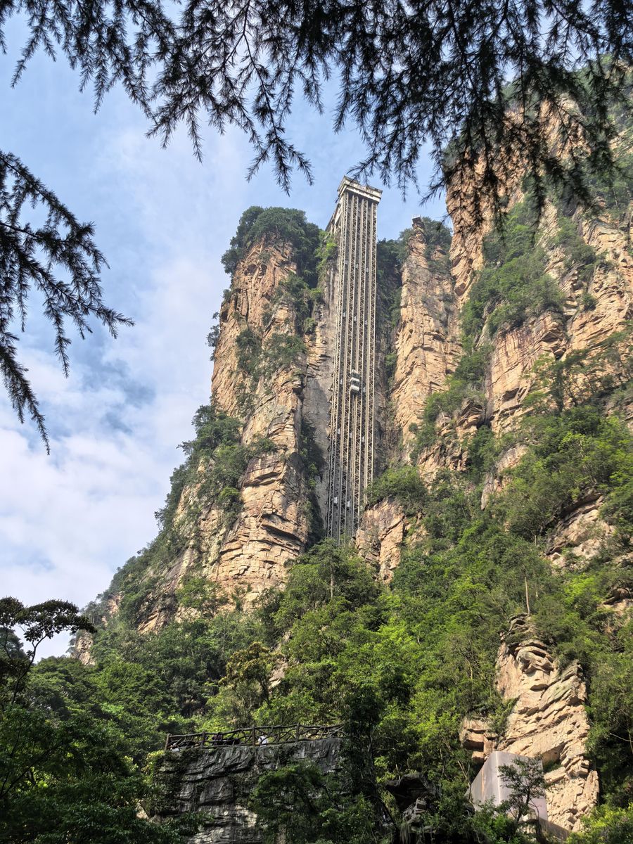 The Bailong Elevator seen from the valley floor: an open steel shaft running up the sandstone cliff, with one glass cabin on its way up, Zhangjiajie National Forest Park