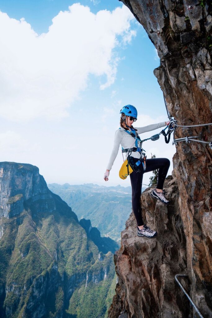 Three climbers on the Sky Ladder of Qixing Mountain seen from below, clipped to the steel cables, with the rest platform on the cliff behind